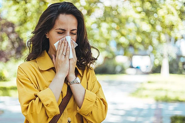 woman with long brown hair wearing a mustard-color blouse and blowing her nose
