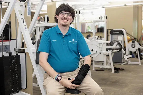 Austin Sisk sitting on a piece of weight equipment in the Atrium Health Floyd Physical Therapy & Rehab Gym