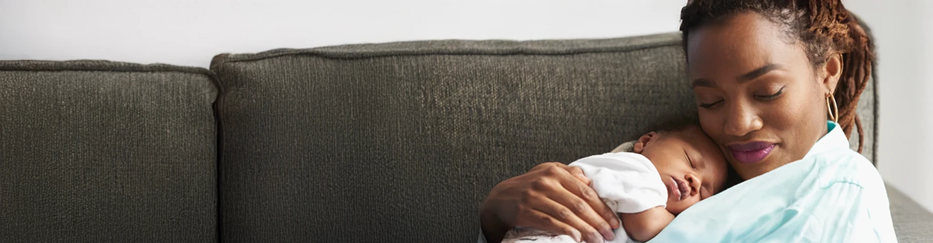A woman sits on her couch with her sleeping newborn baby.