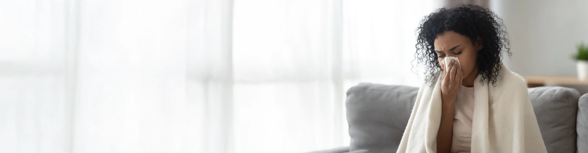 woman with dark curly hair sitting on a gray couch wrapped in a blanket and wiping her nose