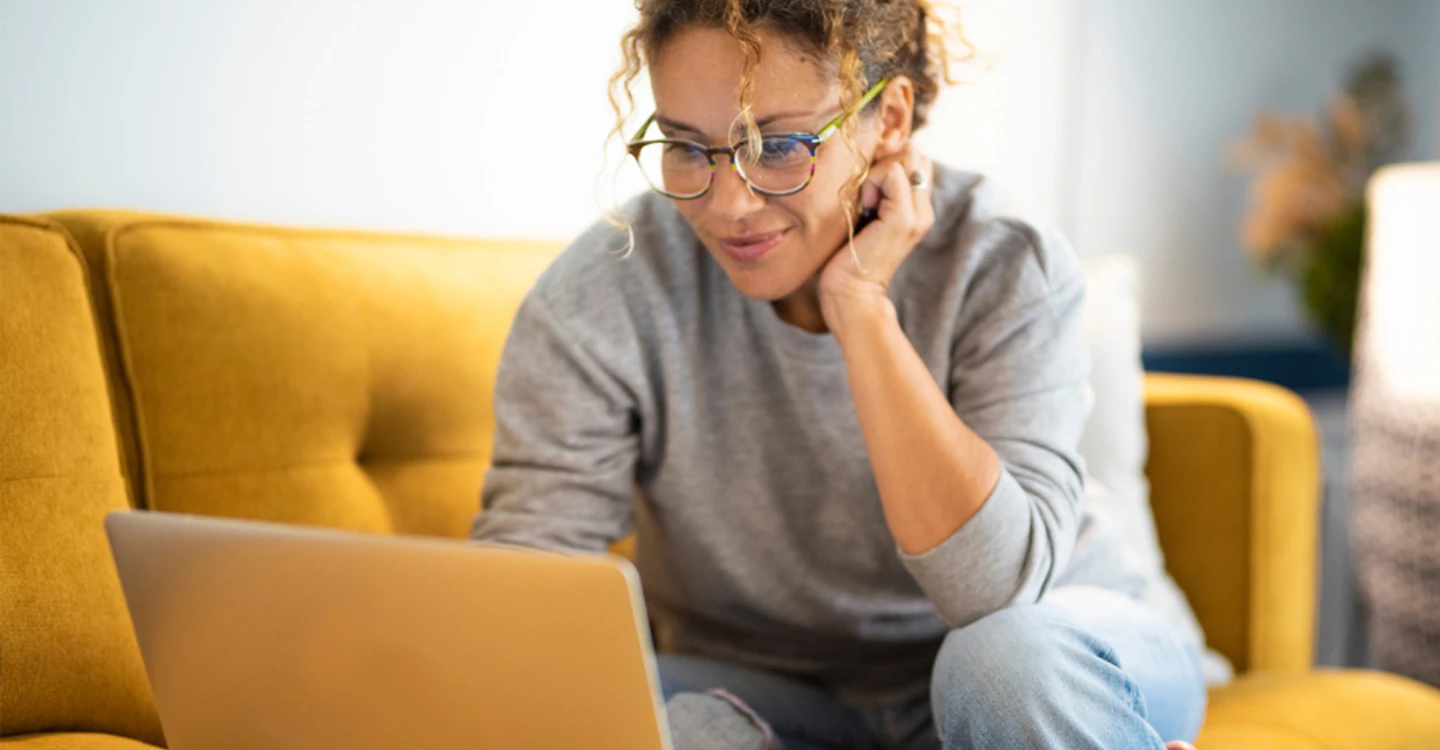 woman wearing glasses sitting on a golden yellow couch and looking at a laptop screen