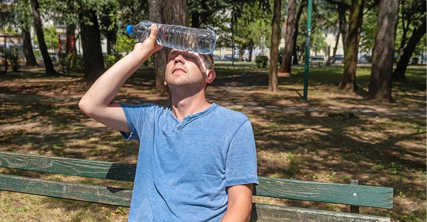 man sitting on a park bench in the sun holding a plastic water bottle on his forehead